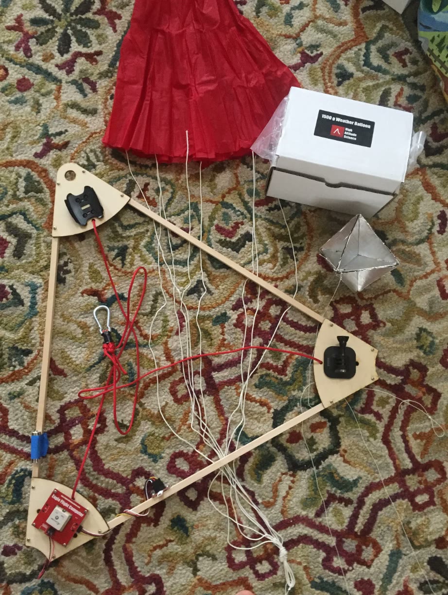 A laser-cut wooden triangle frame with camera mounts, a red parachute, a radar reflector, and a 1,500 gram weather balloon box